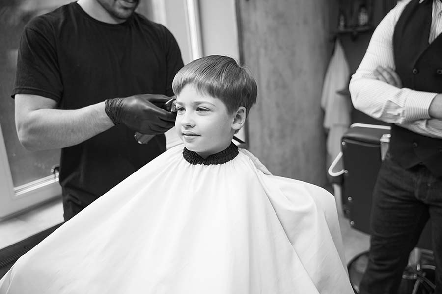 Skilled barber using professional hair trimmer to remove hair from boy's head