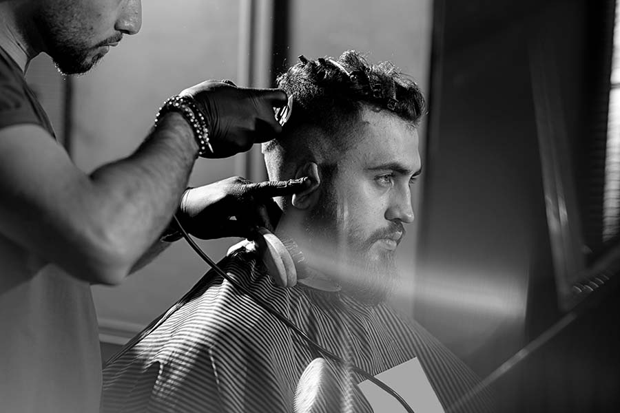 Handsome young man with beard sits at a barber shop. Barber in black gloves shaves hairs at the side.